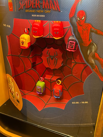 The image depicts a promotional display for a Spider-Man themed McDonald's Happy Meal, likely located in Saalfeld/Saale, Germany. The display features a large cardboard cutout of Spider-Man in his iconic red and blue suit, dynamically posed. In the foreground, arranged on a red, spiderweb-patterned backdrop, are several small, collectible toy backpacks. These toys, designed as mini backpacks, each feature a Spider-Man motif. Some are predominantly red, while others are yellow or green, with varying details and accessories. Text on the display includes "SPIDER-MAN", "BRAND NEW DAY", "NUR IM KINO" (Only in cinemas), and dates "16.07.-01.08." and "02.08. - 19.08.". A portion of a yellow McDonald's promotional stand is visible on the left, with text that appears to be in German, including "Happy Meal" and potentially information about the promotion. The overall presentation suggests a point-of-sale display within a McDonald's restaurant.