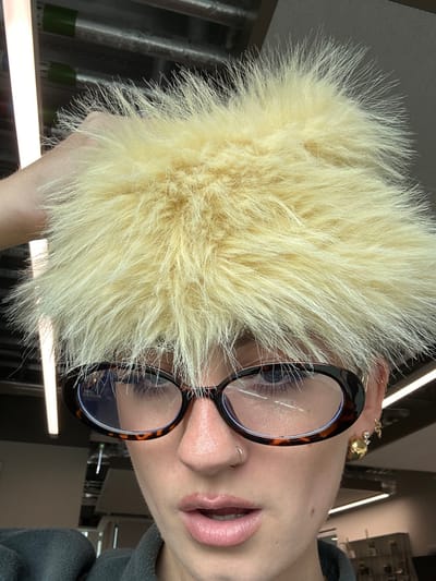 An individual is depicted in a close-up portrait, located in the city of Garston, United Kingdom. The subject's face is centered, viewed from the chest up, with a direct gaze towards the camera. They possess light blonde, spiky hair, portions of which are held upwards by a visible hand on the left side of the head. The individual wears oval-shaped glasses with tortoise-shell frames and clear lenses, revealing light blue eyes. A small silver-colored hoop nose ring is present on the left nostril. The lips are slightly parted. The right ear features multiple piercings, including a gold-colored stud and an additional, smaller earring. The background is an out-of-focus indoor setting, characterized by visible linear fluorescent light fixtures and indistinct shelving or structural elements.