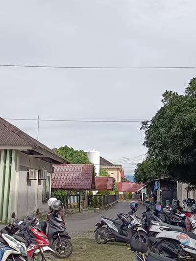A street scene in Mesjid Reuntoh, Indonesia, is depicted under a cloudy sky. The foreground is dominated by a cluster of parked motorcycles, with several more visible further down the street. These scooters, predominantly in white, black, and red, have helmets on their handlebars and seats.
The street itself is paved and appears to be a residential or commercial area, with buildings lining one side. These structures feature red-tiled roofs and light-colored walls, with some exhibiting green vertical paneling. Air conditioning units are visible on the exterior walls of one building.
On the right side of the street, a large, leafy green tree provides a dense canopy. Beyond the buildings and trees, a distant water tower and faint outlines of hills can be seen. Several power lines stretch across the sky, supported by poles. A few individuals are visible in the distance, walking or standing near a structure. The overall impression is of a quiet, daytime scene in a town or village setting.