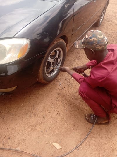 A person wearing a maroon outfit and a camouflage baseball cap is crouching next to the front tire of a black car. They are using a tire inflator to add air to the tire. The car has a clear headlight and a silver rim on the wheel. The ground is a reddish-brown dirt with small pebbles and a fallen leaf. The person is wearing black slides with "NOCTA" written on them. The scene appears to be outdoors during the daytime, with natural lighting. The setting suggests a roadside or a service area in a rural location, consistent with Kwarau, Nigeria. There are no visible emotional cues from the person.