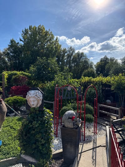 This outdoor image depicts a landscaped garden scene under a bright, sunny sky with scattered white clouds, located in Fretin, France.

In the foreground, on the left, an ivy-covered pillar supports a white classical-style sculpture of a human head, facing slightly right with an open mouth. To its immediate left, a portion of a large dark ceramic urn is visible, and the ground is covered in green vegetation with a partially visible blue object.

Towards the center-right, two red metal garden archways stand over a bed of white gravel, which is bordered by a concrete curb. A dark grey fluted rectangular planter is positioned within the gravel bed, holding a grey, rough-textured sculpture with a small, spherical red object protruding from it. Behind this, green shrubbery is present.

A narrow concrete pathway extends from the midground, running between the gravel bed and a wooden fence with red vertical slats on the right, receding into the background. The background features dense green foliage and numerous trees. The sun is a bright glare in the upper right portion of the sky. No individuals are visible in the scene.