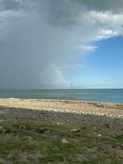 The image depicts a beach scene in Mgudzyrkhwa, Abkhazia. The foreground is dominated by coarse sand and patches of green grass with scattered stones. Beyond this is a shoreline composed of smooth, light-colored pebbles. The sea stretches to the horizon, with waves gently breaking on the shore. The sky is dramatic, with a clear blue section on the right contrasting with dark, heavy storm clouds on the left. A faint rainbow arcs across the darker part of the sky, indicating recent or ongoing rain. No people or structures are visible.