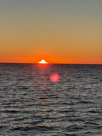A sunset over the Baltic Sea is captured in this image. The sun, a bright white orb with a vibrant orange halo, is partially submerged below the horizon, casting a warm glow across the sky and the water. The sky transitions from a soft blue at the top to a fiery orange near the horizon, creating a dramatic gradient. The sea is a deep, dark blue, with small waves creating a textured surface. Sunlight reflects off the water in streaks of gold and pink, most notably a large, bright pink lens flare in the middle of the frame. There are no people or man-made objects visible in the scene, suggesting a remote or natural setting. The location is identified as Meremõisa, Estonia, a coastal area known for its natural beauty and opportunities to view sunsets over the sea.