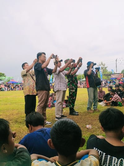 The image shows a group of seven men in a grassy field, located at -7.47 latitude and 111.064 longitude in Karangmalang, Indonesia. Six of the men are looking at something in the distance, and they are all holding up their mobile phones, appearing to take pictures. The seventh man is standing next to the others, but he is looking at the camera, and is also holding a mobile phone up to take a picture. The men are wearing a variety of clothing, including casual shirts and pants, and a few of them are wearing hats. The environment is outdoors in a grassy area with a few trees in the background. There are also other people visible in the background, as well as a blue banner. There are a few kids sitting in the foreground, looking in the same direction as the men, with their backs to the camera. The scene appears to be at a public event of some sort, with many people present.
