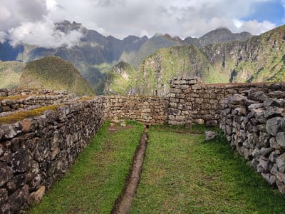 Stone walls enclose a grassy area, possibly ruins, with a narrow dirt trench running through the grass. In the background, lush green mountains rise under a cloudy sky. A faint rainbow is visible above the mountain peaks. The stone walls are weathered, with some sections covered in moss. The area appears to be part of an ancient construction.