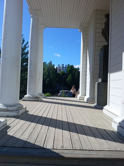 The image depicts an outdoor scene viewed from a covered porch with white columns and a wooden floor. In the distance, through an opening in the porch, a ruined castle-like structure is visible, surrounded by lush green trees under a bright blue sky with scattered clouds. The sun appears to be high in the sky, casting distinct shadows across the wooden planks of the porch, suggesting it is midday on a clear day. A person with blonde hair, wearing a light-colored top, is seated on the porch, looking towards the distant castle. The architecture of the porch, with its classical columns and wooden detailing, hints at a historical or scenic location. The overall mood is peaceful and serene, with the natural beauty of the landscape contrasting with the man-made structures. The setting appears to be Vyborg, Russia, as indicated by the location context.