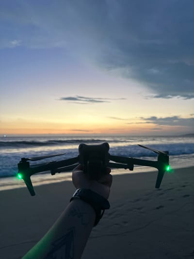 A person holds a drone with green lights on the beach at Brisas de Zicatela, Mexico. The beach and the ocean are behind the drone. The sky is visible. The person's arm has a tattoo on it. The person is also wearing a watch.