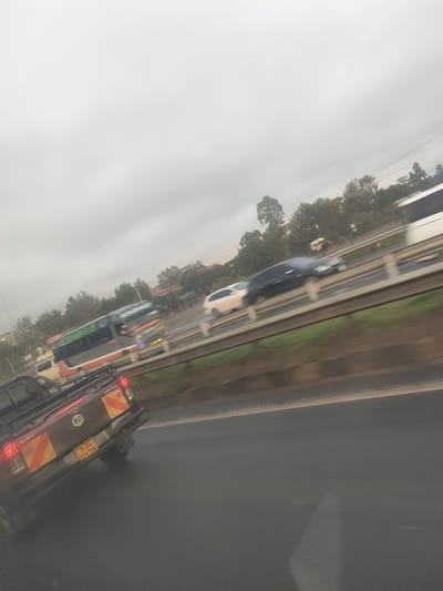 The image depicts a multi-lane highway scene in Biashara ward, Kenya, under an overcast sky. In the foreground, the rear of a brown pickup truck with a `KDC 017M` Kenyan license plate is visible, occupying the left lane and moving away from the viewer. The truck features reflective orange and yellow chevron markings on its tailgate and rear bumper, along with a circular "80" speed limit decal. To the right of the truck is a dark asphalt road surface. In the mid-ground, an elevated section of a multi-lane highway is separated by a metal guardrail and a vegetated median. Multiple vehicles on this elevated section exhibit motion blur, indicating movement. These include, from left to right: a light-colored vehicle (partially visible), a grey and green intercity bus, a white sedan, a dark-colored SUV, and a white, possibly large, vehicle (partially visible). The vehicles are in motion, traveling in the opposite direction to the foreground pickup truck relative to the camera's perspective. The background features a dense line of green trees, with indistinct structures partially visible among them. The sky is uniformly grey and overcast. No individuals are clearly discernible.