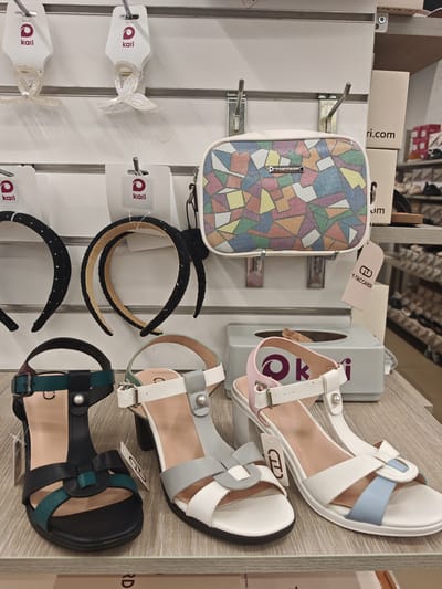 This media file is a close-up, eye-level shot of a retail display featuring women's accessories and shoes. The scene is set within a store, likely a shoe or fashion boutique, indicated by the organized display shelves and branded packaging.
The primary subjects are various fashion items. On the upper shelves, there are clear plastic hair clips with iridescent finishes, each attached to white cardboard tags bearing the "Kari" logo. Below these, two black headbands, one adorned with small rhinestones and the other with a textured beaded pattern, are displayed.
In the center of the frame, a small, rectangular handbag with a colorful geometric mosaic pattern is showcased. It hangs from a metal hook. Adjacent to the handbag, a light gray tissue box is visible, with a pink and white sandal strap extending over it.
The lower shelves display three pairs of women's sandals. The leftmost sandal is black with teal accents and a T-strap design. The middle sandal is white with light gray and white straps. The rightmost sandal is white with light blue and white accents. All shoes are presented on a light wood-grain surface.
Behind the handbag and tissue box, white cardboard boxes are stacked, one of which has "ri.com" printed on it. A pink tag with a "T-TACCARDI" logo is also visible. The lighting appears to be artificial, typical of an indoor retail environment, with no indications of time of day or weather. The overall impression is one of commercial display, focusing on the presentation of merchandise.