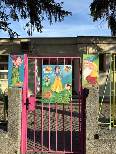 A bright pink metal gate is positioned in front of a light-colored building. On the gate, and attached to concrete pillars on either side, are colorful painted panels depicting characters from the fairy tale "Snow White and the Seven Dwarfs." On the left pillar, a dwarf in a green tunic and hat is painted. On the gate itself, a panel shows Snow White in her yellow dress, with Winnie the Pooh and a bell above, a little girl with a red hood, and a squirrel. On the right pillar, another dwarf, this one with a long white beard and a blue hat, is painted. The building behind the gate has an air conditioning unit mounted on its facade. The ground in front of the gate is paved with concrete slabs, and there are shadows cast by the gate and nearby trees. The scene appears to be outdoors, under a blue sky with some wispy clouds and branches of a coniferous tree hanging from the top of the frame. The location is identified as Kaspichan, Bulgaria.