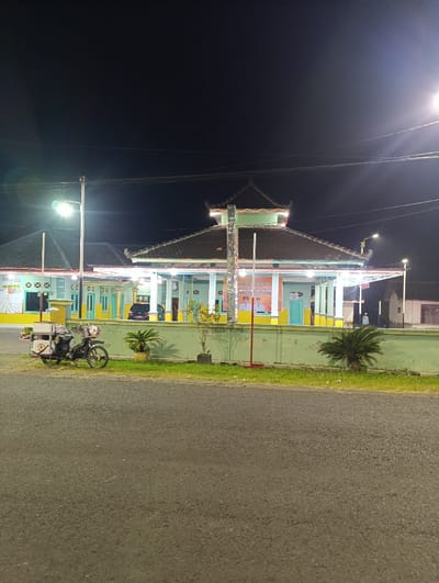 The image shows a brightly lit, traditional building in the Kecamatan Kapongan, Indonesia (latitude: -7.675, longitude: 114.065), at night. The building has a tiled roof with a  wooden frame and a  light green exterior wall. It features a covered veranda with light fixtures. There are two palm trees in front of the building with one potted plant on each side. Behind the building is a low wall that has a light red painted line.  In the front of the building, there is a small motor scooter parked near a low wall. There are several streetlights illuminating the building. The building looks like a community center or a place of worship.  There is a person standing outside the building to the left of the image, who looks like they are dressed in a white long-sleeved shirt.