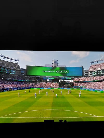The image shows a professional soccer match being played in a stadium. The stadium has a large green scoreboard displaying "POWERED BY aramco". The field is well-maintained, with visible white lines marking the boundaries and center circle. There are players on the field, appearing to be in the middle of a game, with spectators filling the stands in the background. The overall impression is a lively sporting event under a partly cloudy sky. The image does not provide any information about the city of Altea, Spain.