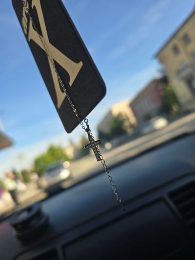 The image presents a close-up, angled view from inside a vehicle, looking outward through a windshield. The primary subjects are decorative items hanging within the car's interior. A thin, silver-colored chain extends downwards, attached to a small, ornate cross pendant. The cross is adorned with multiple small, sparkling stones or crystals.

The chain further connects to a black, rectangular object, which appears to be made of a matte material. This object prominently displays a large, stylized white letter "X" on its surface. The overall appearance of the black object and its hanging nature suggests it could be a car air freshener or a similar accessory typically suspended from a rearview mirror.

In the heavily blurred background, visible through the vehicle's window, there is a clear, bright blue sky, indicating daytime. Below the sky, indistinct forms of light-colored buildings and some green foliage can be seen, suggesting an urban or suburban environment. The scene is static, depicting no active event. The time of day is clearly daylight, with clear weather conditions. No people are visible. The location context for this scene is Byala, Bulgaria. The visible text is the large white "X" on the black rectangular object.