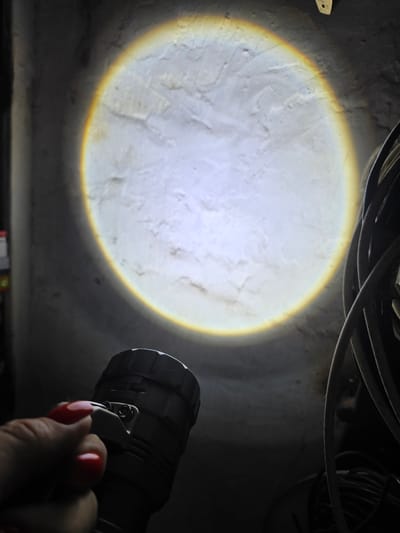 An image captures a hand with red nail polish holding a dark-colored flashlight, directed upwards. The flashlight beam illuminates a textured, off-white wall, creating a large, circular spotlight with a subtle yellow-orange halo around its edge. To the right of the wall, thick, dark electrical cables are bundled together. The scene is dimly lit, with the wall and cables partially obscured by shadows. The image does not contain any contextual information related to the city of Nova Kakhovka, Ukraine.