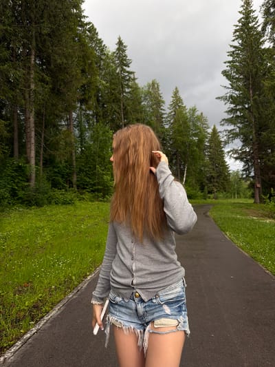 A young woman with long, light brown hair is standing on a paved path in a park-like setting. She is wearing a grey cardigan and ripped denim shorts. Her hair is falling over her shoulders and she is touching her hair with her right hand. She is holding a white phone in her left hand. The path is bordered by a grassy area with small white flowers and tall green trees. The sky is overcast, suggesting a cloudy day. The trees appear to be pine and other coniferous varieties, typical of a northern climate. The overall scene depicts a casual moment outdoors, possibly during a walk or break in a park. No text is visible in the image. The location context is Chusovoy, Russia.