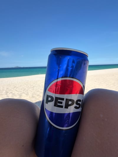 A close-up shot features a Pepsi can held by a person's legs on a sunny beach. The can, partially obscured by the legs, displays the iconic Pepsi logo. In the background, a pristine white sandy beach meets the turquoise ocean under a clear blue sky. In the distance, a small island is visible. The scene suggests a relaxed day at the beach in a tropical or coastal location, possibly Bac Cam Ranh Ward, Vietnam, given the context.