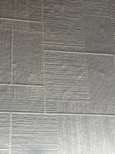 The image shows a close-up, overhead view of a textured surface composed of rectangular and square panels arranged in a grid-like pattern. The surface appears to be flooring or a wall covering with a matte finish and a color resembling dark grey or charcoal. Each panel features a distinct linear texture, with some panels having vertical lines and others having horizontal lines. The lines are raised, creating a tactile and visually interesting surface. The panels are separated by thin, lighter-colored grout lines, which are also arranged in a perpendicular grid. The lighting in the image casts subtle shadows that highlight the texture of the surface. There are no people, animals, or distinct objects visible in the frame. The context of Yuzhno-Sakhalinsk, Russia, is not evident from the visual information provided in the image itself, as the scene is an abstract textured surface without any identifiable landmarks or environmental cues specific to any location.