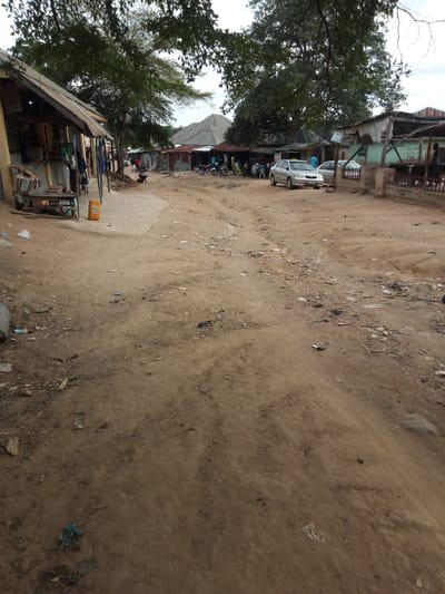 The image depicts a dirt road in what appears to be a semi-urban or rural area. On the left side of the road is a low-lying structure with a corrugated metal roof, resembling a shop or market stall. Several people are visible near the stall, and some items, including what looks like eggs in a carton, are displayed on a wooden table outside the structure. A yellow plastic container is also present. Further down the road, a silver sedan car is parked, and another vehicle, possibly a motorcycle, is visible in the distance. The road itself is unpaved and shows signs of wear and tear, with scattered debris, including plastic bags, paper, and other refuse, along its surface. The environment is characterized by dusty soil, sparse vegetation, and a cloudy sky. Buildings with metal roofing are visible on both sides of the road, suggesting a settlement or village. The overall impression is one of a functional, albeit unkempt, community pathway.