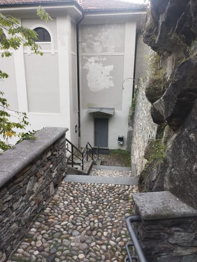 The image shows an exterior view of a narrow stone staircase descending from a wall. The staircase is bordered by a stone wall on the left and a rocky incline on the right. The steps are made of cobbles and concrete. At the bottom of the stairs, there is a gray door and a white building with a textured wall. A streetlamp with a curved arm is visible attached to the side of the building. In the foreground, a stone railing with a metal handrail runs alongside the stairs. Green leaves from a tree are visible in the upper left corner. The scene appears to be located in Orselina, Switzerland, likely a historical area with a mix of stone architecture and natural rock formations. There are no people or active events depicted in the image.