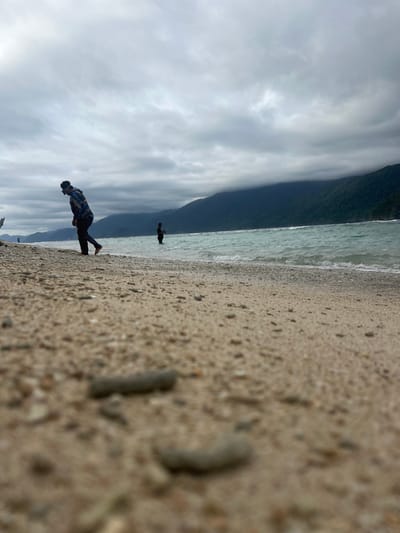 This is a vertical, eye-level shot of a beach in Babahie, Indonesia. The foreground is dominated by a sandy beach with visible pebbles and some coral fragments. To the left of the frame, the sand extends out of focus. To the right, the clear, shallow water of the ocean laps gently at the shore, with small waves visible. 

In the mid-ground, a person stands in the shallow water, facing away from the camera. They are wearing dark clothing and appear to be walking or standing. Further back, on the beach to the left, another person is standing, also facing away. This person is dressed in a dark, patterned jacket, dark pants, and shoes. They appear to be looking out at the ocean or the horizon. 

The background features a lush, green mountainous landscape that meets the water. The sky above is overcast with heavy, white clouds, suggesting it might be a cloudy day or nearing sunset. The overall mood is serene and peaceful. There is no visible text in the image.