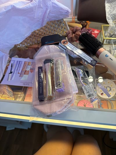 This close-up, top-down shot captures a collection of beauty and hair care products neatly arranged within what appears to be a vanity drawer or organizer, likely indoors in Groningen, Netherlands. On the far right, part of a person's bare thigh and leg are visible, suggesting human presence nearby.
The central subject is a light pink, ribbed corduroy pouch containing several items still sealed in plastic. Above and to the left of the pouch, a prominent white and rose gold hot air brush with black bristles features an "AUTO" setting. Below the brush, multiple scrunchies in shades of brown and beige are visible.
Other notable objects include a box for "lights, camera, lashes platinum mascara" and a lavender-packaged "shape tape blur concealer brush," both from the brand "tarte." A daisy-patterned compact mirror with a ladybug sticker and a pink compact are also seen. A promotional leaflet or catalog for "tarte high-performance naturals" displaying various makeup products lies beneath the pouch.
Visible text includes product names and descriptions such as "MARACUJA JUICY LIP PLUMP," "ULTRA creamy," "24-hour moisturizing," "hydratation 24 heures," "fae-light neutral," "shape tape blur concealer brush," "pinceau anti-cerne," "lights, camera, lashes platinum mascara," and "mascara platinum." The brand name "tarte" is frequently visible across multiple items. Other text like "FG15479-UN," "brush shot," and "co-anda2x" are also present. The scene suggests an organized collection of new or recently acquired beauty and personal care items.