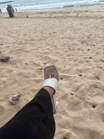 The image shows a person's legs and feet standing on a sandy beach. The person is wearing black pants and white slide sandals. The sand is light brown and has footprints and small pebbles scattered on it. In the background, the ocean water is visible, appearing calm and light blue. The shoreline is wet, suggesting the tide is close. Further in the distance, the waves are breaking gently. The lighting suggests it could be daytime, possibly overcast or early morning/late afternoon given the diffused light. There are no discernible emotions conveyed by the image. The setting is clearly a beach, with the sand and ocean being prominent elements. No text is visible in the image.