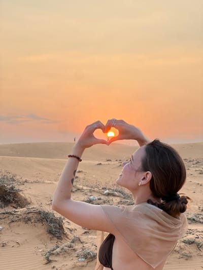 A forensic description of the image reveals a female subject posing against a sand dune landscape at sunset, identified as the Red Sand Dunes near Phan Thiet, Vietnam.
**Scene Description:**
The primary subject is a female, captured from the upper torso upwards, positioned in profile facing left. Her dark brown hair is pulled back into a low bun, secured with a black elastic. She exhibits a smiling expression, looking upwards and to the left. She wears a dark brown or black bikini top, partially covered by a sheer, shimmering light beige fabric draped over her shoulders and upper back. A small, light-colored stud earring is visible in her left ear.
Her arms are raised, with both hands forming a heart shape directly in front of the low-lying sun. The sun, a bright orange orb, is centered within the heart. On her left wrist, a multi-bead brown bracelet is present. A dark, circular or abstract design, possibly a tattoo, is visible on her upper left arm. Her fingers are interlocked to create the heart form. Multiple silver-toned rings adorn her fingers: on the left hand, rings are present on the middle and ring fingers; on the right hand, an X-shaped ring is prominent on the middle finger, with another ring on the ring finger.
The background consists of extensive light brown/tan sand dunes, characteristic of the Red Sand Dunes. Sparse, dry vegetation is scattered across the foreground and midground dunes. In the mid-distance, silhouetted against the bright sky atop a distant dune, a faint human figure is discernible. The sky displays a gradient of warm hues, transitioning from a brilliant orange-yellow at the horizon, indicative of sunset, to a softer peach and light orange higher up. White debris is visible among the foreground vegetation.