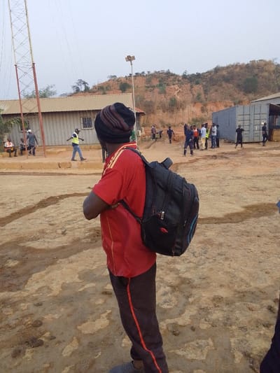 The image depicts an outdoor scene in Hoche, Nigeria, characterized by an unpaved, reddish-brown dirt ground with visible depressions and uneven patches. In the immediate foreground, facing away from the camera and slightly to the right, is a person wearing a red short-sleeved shirt with yellow shoulder accents, dark trousers with a red stripe, and a striped knit beanie (black, brown, red). This individual carries a black backpack with blue accents.

The midground features various structures and groups of people under an overcast sky. To the left, a tall metal lattice tower stands beside a single-story building with a light-colored corrugated metal roof and walls, equipped with barred windows. Several individuals are present near this building; one person in a light safety vest walks from left to right, while others stand or sit. Centered in the midground, a wooden pole topped with a solar panel and light fixture is visible. Further to the right, a cluster of approximately ten individuals, some appearing to wear work attire or safety vests, stand and interact. Behind them, multiple grey shipping containers are utilized as structures, with one appearing partially open. Another structure with a corrugated metal roof is partially visible on the far right.

The background consists of a prominent reddish-brown hill with sparse, dry vegetation, suggesting a semi-arid or excavated landscape. The overall environment appears to be an active work site or a transitional area.