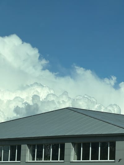 The image shows a building under a blue sky with voluminous cumulus clouds. The building's exterior appears to be made of gray panels and features vertical rows of windows with white frames. Reflections of trees or other foliage are visible in the windows. The exact location cannot be verified based solely on the image and without additional context or identifying information about Vereya, Russia.