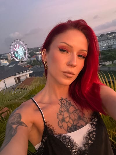 A woman with bright red hair is in the foreground, wearing a white top with black lace trim. She has a large floral tattoo on her chest and another tattoo on her left arm. She has gold-colored earrings and a piercing in her nose. Behind her, the Ferris wheel is visible, a landmark that identifies the city of Tibubeneng, Indonesia. The environment includes city buildings, palm leaves, and a gray sky, possibly at dusk or dawn.