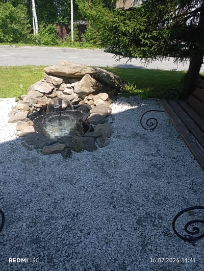 The image depicts an outdoor scene with a small rock garden featuring a water fountain. The fountain is centrally located within a circular arrangement of stones, with water spraying upwards. Surrounding the fountain is a bed of white gravel. To the right, a wooden bench with decorative black metal supports is partially visible, positioned on the gravel. In the background, lush green trees and foliage are present, along with a paved surface, likely a road or pathway. The lighting suggests it is daytime with shadows cast on the ground. The timestamp at the bottom right indicates the image was taken on July 16, 2026, at 14:41. The text "REDMI 13C" is also visible in the bottom left corner. This scene is likely a garden or park area.