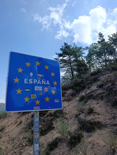 A rectangular blue sign with a border of yellow stars, representative of the European Union, displays the word "ESPAÑA" in white lettering. Several smaller stickers are affixed to the sign. The sign is mounted on a metal pole which is covered in additional stickers. The sign is situated on a dry, rocky, and overgrown hillside under a bright blue sky with scattered white clouds. Green trees are visible in the background. The location is Água da Pala, Portugal.