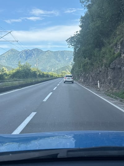 The image captures a view from a vehicle traveling on a paved road that runs alongside a steep, rocky mountainside covered in dense green foliage. A white van is visible ahead on the road. The road itself is marked with white dashed lines. To the left of the road, the terrain slopes upwards towards the mountain. In the distance, more mountains can be seen under a bright blue sky with scattered white clouds. Power lines with insulators are also present in the landscape. The image is oriented such that the road and the scene appear to be running vertically. The lighting suggests it is daytime, likely late morning or afternoon, with clear weather. The overall impression is one of travel through a scenic, mountainous region.
