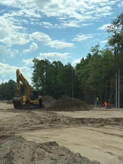 This image depicts a construction site in Kabanikha, Russia, on a partly cloudy day. A yellow Komatsu excavator is parked on sandy ground, with its bucket lowered. A construction worker in dark clothing stands beside the excavator. In the background, a large pile of dirt is visible, along with a line of trees. Further to the right, another construction worker in an orange safety vest and pants is operating surveying equipment. A tripod with a green prism attachment stands next to him. Electrical poles are also present on the right side of the image. The ground is uneven, with tire tracks from heavy machinery evident in the sand.