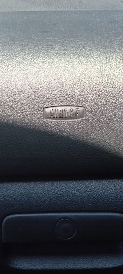 The image shows a close-up view of an "AIRBAG" label embossed on a textured surface, likely the dashboard of a vehicle. Below the label, there is a darker, horizontal panel with a recessed area that appears to be a compartment or a handle. The lighting suggests an interior setting, with a shadow cast across the upper portion of the airbag label. The scene is forensic and matter-of-fact, focusing solely on the visual elements present in the image. No location in Koviahy, Ukraine, is discernible from the image itself, as it depicts a generic car interior.