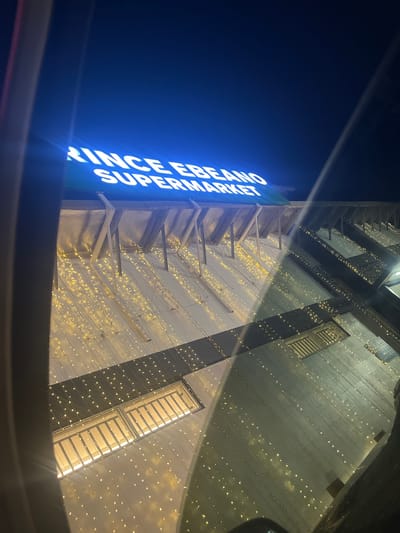A nighttime exterior shot captures the brightly lit sign of "PRINCE EBEANO SUPERMARKET" in Ojomu, Nigeria. The building is adorned with numerous strings of small, warm-toned lights that cascade down its facade, creating a festive and inviting atmosphere. Above the lights, the building's structure is visible, with angled supports and a reflective material on its upper sections. Two rectangular windows with illuminated interiors are prominent on the lower portion of the building, suggesting interior activity. The image is taken from a slightly elevated angle, with dark structures in the foreground partially obscuring the view. The sky above is a deep, dark blue.