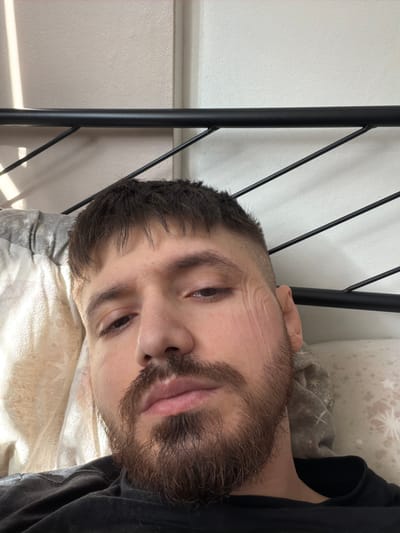 A close-up, slightly skewed portrait shows a light-skinned man with dark hair and a full, short beard. He appears to be lying on a bed with a patterned comforter and a portion of a black metal bed frame is visible in the upper left. He is wearing a dark-colored shirt. No elements in the image suggest it was taken in Pentre, United Kingdom.