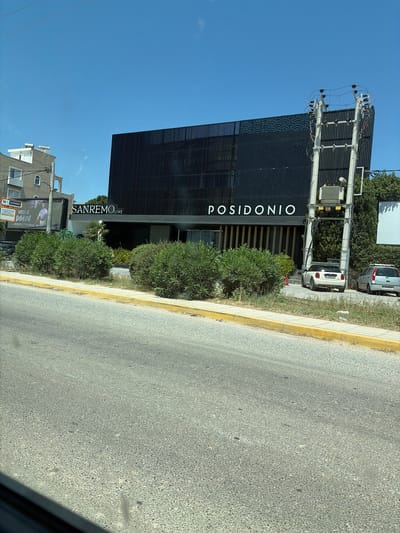 The image shows the exterior of a modern building with the name "POSIDONIO" prominently displayed in large white letters. Below this, the words "SANREMO LIVE" are also visible. The building has a dark, slatted facade and is surrounded by lush green bushes. To the left of the building, there's a street with a yellow curb and pavement, suggesting an urban or commercial setting. A few cars are parked near the building, and trees are visible in the background, some of which appear to be part of the natural landscape. The sky is clear and blue, indicating a sunny day. The perspective suggests the photo was taken from a moving vehicle, possibly looking slightly upwards at the building. The overall scene appears to be a daytime view in a developed area, possibly a venue or establishment.