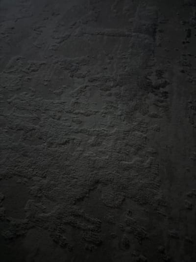 The image displays a close-up, dimly lit view of a textured, dark gray surface. The texture appears to be rough and uneven, with raised ridges and depressions that create shadows and highlights. The material could be concrete, stone, or a similar rough-textured substance. There are no people, objects, or discernible landmarks visible in the image that would indicate its location within the city of Tomsk, Russia. The perspective is angled, suggesting the surface is either a wall, floor, or a large, irregular object.