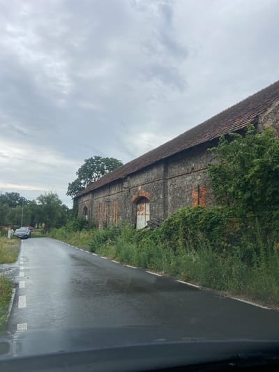 The image shows an old, weathered stone building with a red tile roof, situated in Kruszyn, Poland. The building appears to be a long barn or storage facility, with arched openings and visible brickwork. The exterior walls are constructed from large, irregularly shaped stones, interspersed with vertical bands of red brick. Several arched doorways are visible, one of which has a rusted metal door. The building is overgrown with lush green vegetation, including ivy climbing the walls and tall grasses growing around its base.

To the left of the building, a wet asphalt road curves away from the viewer. The road has white dashed lines marking the lanes. A dark colored car is parked on the left side of the road. The surrounding environment is natural, with mature trees visible in the background, and a cloudy sky overhead, suggesting overcast weather. The overall impression is one of age and disuse, with nature reclaiming the structure.