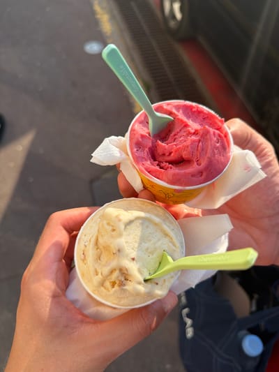 Two hands hold two small cups of gelato outdoors in Saint-Tropez, France. The cup in the upper portion of the image contains pink gelato, likely strawberry or raspberry, with a green, compostable spoon embedded in it. The spoon has "Compostable" printed on it in white text. A white paper napkin is visible around the rim of this cup. The cup in the lower portion of the image contains pale yellow gelato with visible chunks of nuts or fruit, also with a green, compostable spoon. The background shows a paved surface and a dark object, possibly a car or building. The lighting suggests it is daytime, with shadows indicating sunlight.