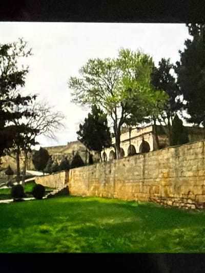 The image shows a stone wall bordering a lush green lawn in front of an ancient-looking structure with arched openings. Several trees are visible, some with vibrant green foliage and others bare-branched, suggesting a temperate climate. The sky above is overcast. The architecture of the building, with its stone construction and arched facade, hints at historical significance, possibly a palace or a religious site. The overall scene evokes a sense of tranquility and historical depth. However, there is no information provided in the image itself or the crops that allows for specific identification of the location within Lopatino, Russia.