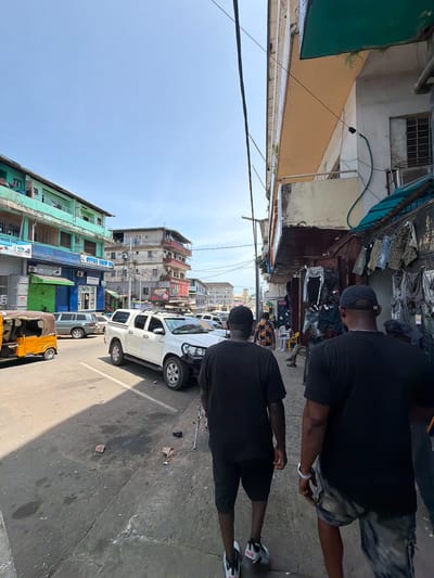 The image depicts a bustling street scene in a city, consistent with Monrovia, Liberia, viewed from street level. The perspective is looking down a narrow thoroughfare lined with buildings and activity.

In the foreground, two men are walking away from the camera. The man on the left wears a black t-shirt, patterned shorts, and a silver-colored bracelet. The man on the right is dressed in a black t-shirt, black shorts, and white and black athletic shoes with red accents. Further back, other individuals can be seen amidst vendor stalls.

The street is paved, showing signs of wear and some damp spots. Vehicles include a white pickup truck parked on the right side and a yellow auto-rickshaw (tuk-tuk) further down the street.

The buildings on both sides of the street are multi-story, exhibiting various architectural styles and states of repair. Colors like green, beige, and white are prominent. Many buildings appear somewhat aged, with some showing exposed rebar. Commercial establishments line the street, with goods, possibly clothing, displayed at outdoor stalls on the left. Overhead utility lines are visible against the sky.

Visible text on building signs includes "OFFICE EQUIP INC", "TECH ENTERPRISES", "HUB INC", "Canon", and "GLUCKST". The bright lighting and clear sky indicate it is daytime. The overall scene suggests a vibrant, active urban environment.