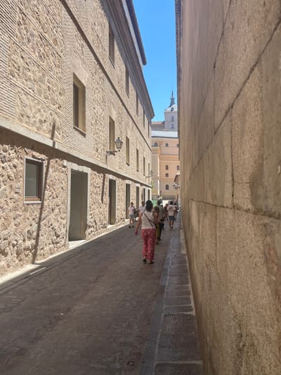 The image depicts a narrow, cobblestone street in Toledo, Spain, with tall, stone buildings flanking both sides. The street is cast in shadow, with sunlight illuminating the upper portions of the buildings and the sky above. Several people are walking down the street, appearing to be tourists. On the left, a building exhibits a facade of alternating stone and brickwork, with several windows and doorways visible. A decorative streetlamp is attached to the wall. On the right, a massive stone wall dominates the view, its rough texture and cracks evident. In the distance, at the end of the street, a prominent tower with a spire rises against the clear blue sky, suggesting a significant architectural landmark within the city, likely a church or cathedral. The overall impression is one of historical charm and a warm, sunny day in a European city.