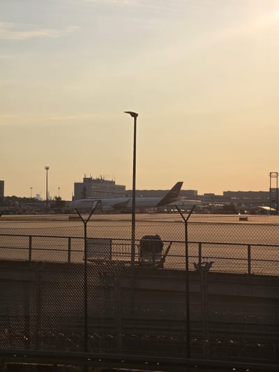 The image depicts an outdoor scene at the Frankfurt Airport in Kelsterbach, Germany, during what appears to be late afternoon or early evening, indicated by the warm, golden light and long shadows. In the foreground, a chain-link fence runs horizontally across the lower portion of the frame, with barbed wire visible below it. Behind the fence, a tarmac area is visible, with a large Lufthansa airplane parked in the middle ground. The airplane is predominantly white with the airline's logo on its tail. Further in the background, several airport buildings and structures, including what appears to be a control tower, are silhouetted against the sky. A lamppost stands vertically in the right middle ground. The sky is a pale yellow-orange, with some wispy clouds. The overall impression is one of an operational airport at the end of a day.