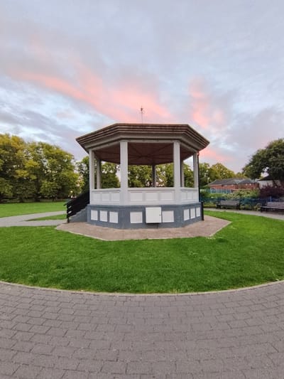 The image shows a hexagonal bandstand in a park. The bandstand is white with grey accents and has an open structure supported by pillars. A set of dark stairs leads up to the bandstand. The bandstand is surrounded by a well-maintained green lawn. A paved pathway curves around the lawn in the foreground. In the background, there are trees, benches, and buildings. The sky is visible above, with soft pink clouds suggesting either sunrise or sunset. The scene appears to be peaceful and serene. The bandstand is located in the town of Evesham, within the Wychavon district of Worcestershire, United Kingdom.