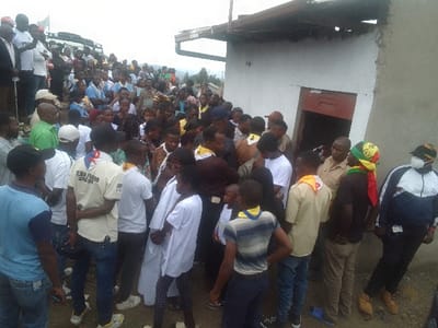 A large crowd of people stands outside a simple, one-story building with a partially visible red door. The diverse group includes individuals in casual clothing, uniforms, and some with scarves. Some wear scout neckerchiefs. The crowd appears dense, with people of various ages and skin tones intermingled. Overlooking the building, the landscape gently slopes downward, providing a backdrop of distant trees and cloudy sky.