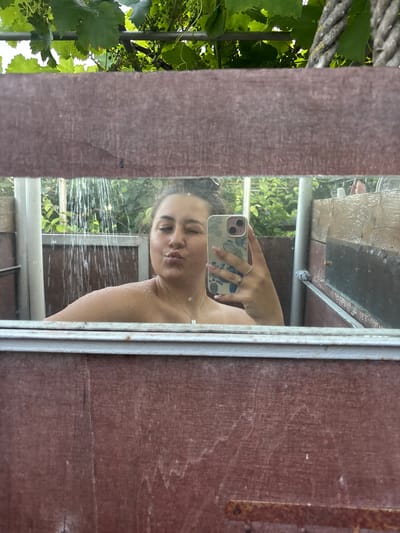 This media file captures a selfie taken in a mirror, showing a young woman in what appears to be an outdoor shower. The woman is leaning in towards the mirror, puckering her lips as she takes the photo with her phone, which has a floral case. She is wearing a necklace. Water is spraying down, visible as streaks in front of her and on her skin, indicating she is showering.

The setting seems to be outdoors, with lush green foliage, possibly vines, visible in the background through the shower. There is a rustic wooden structure framing the mirror, and hints of metal railings. The overall impression is of a casual, perhaps vacation-like, setting. The lighting suggests it's daytime, possibly a sunny day given the bright greenery. The woman's expression is playful and relaxed, suggesting enjoyment of the moment. There are no visible text elements in the image.