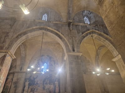 The image shows the interior of the Crusader Church of Saint Abraham in Abu Gosh, Israel. The scene is dimly lit, with a strong sunbeam emanating from the right side, illuminating dust particles in the air. The architecture features high, arched ceilings and stone walls, consistent with medieval construction. On the left wall, faded frescoes depict religious scenes and figures. Several modern chandeliers hang from the ceiling, providing additional light. Small, patterned windows are visible in the upper walls. The overall impression is one of an ancient religious site with some modern interventions.