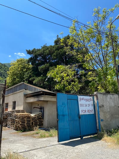 The image captures an outdoor scene on a clear, sunny day in Mtskheta, Georgia. The foreground features a concrete driveway with scattered patches of dry grass. To the left, a stack of wooden pallets is piled high against a tan-colored building. This building has a corrugated metal roof and several barred windows. Adjacent to the building is a tall, blue metal gate, partially open, revealing a glimpse of what lies beyond. Attached to the gate is a white sign with red and black text in Georgian and English, indicating "FOR RENT" and a phone number.
Behind the gate and to the right, a concrete wall borders the property. Overhanging the wall and extending into the frame are lush green trees, with one large pine tree dominating the background. In the far distance, a verdant, mountainous landscape is visible under a bright blue sky with a few wispy white clouds. Power lines crisscross the upper portion of the image. The overall impression is one of an industrial or storage area in a semi-rural or suburban part of Mtskheta.
